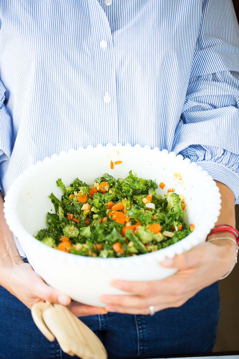 Woman holding a white salad bowl filled with greeans and carrots and other fruits and vegetables.