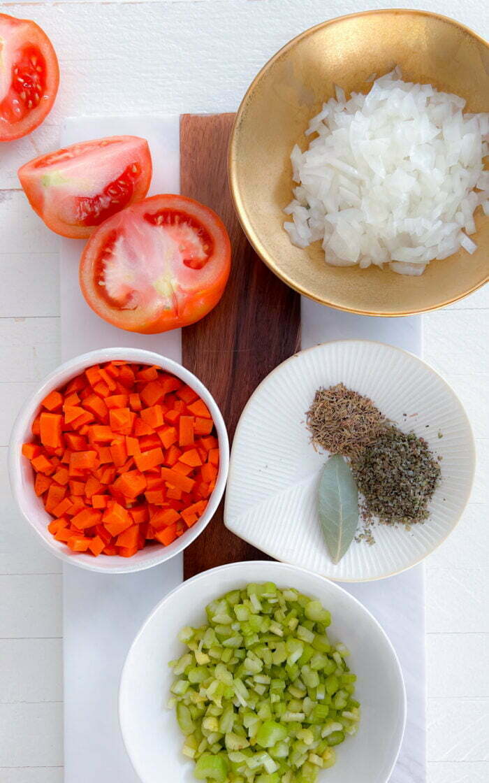 Diced oinon in a gold bowl, chopped carrots and celery in a white bowl, spices in a white plate.