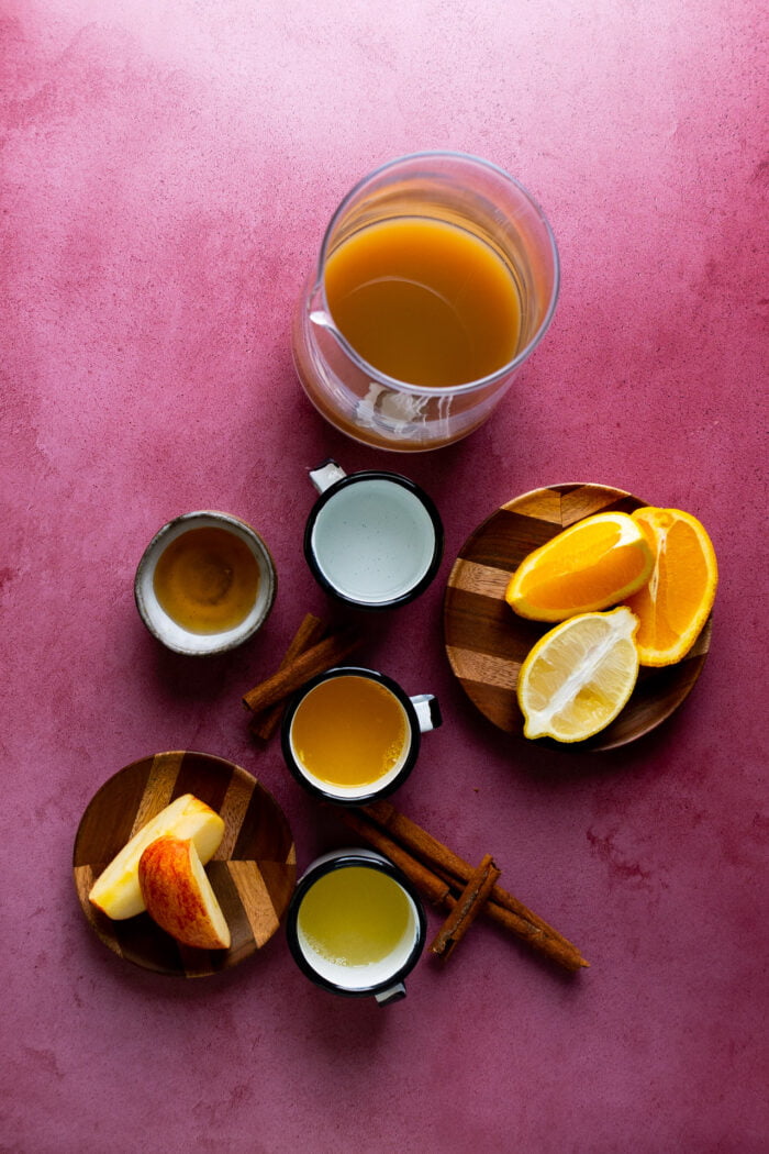 apple cider in a glass pitcher, agave syrup, orange juice and lemon juice nesxt to sliced apples and oranges.