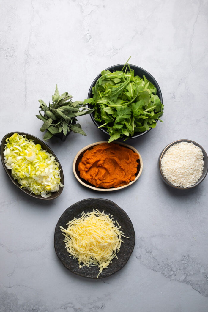 Ingredientes para hacer risotto de calabaza vegano. Arúgula, arroz arborio, queso parmesano, puré de calabaza y rodajas de puerro.
